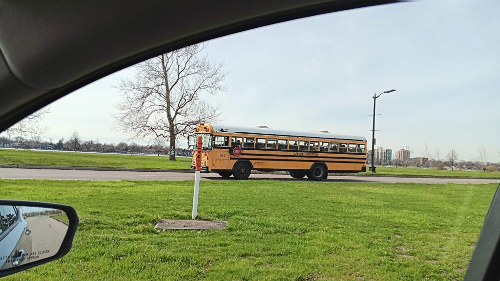 A yellow school bus drives students from the James and Grace Lee Boggs School to a field trip on Belle Isle in Detroit Michigan. There is a bright green lawn and the photo is being taken from a car window. 