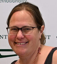 Portrait style photo of SEMIS education specialist Sarah Halson wearing glasses and smiling at the camera with a shoreline behind her.