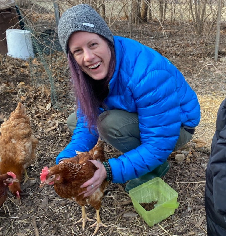 A white woman with purple hair is crouching down with her hands on either side of an orange chicken. There is a green plastic bin on her left and two more chickens on the right side. She is wearing a gray beanie, a bright blue puffy jacket, green pants, and is smiling.