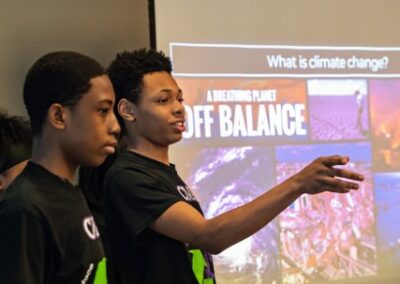 Three students in black shirts do a presentation on climate change they are standing in front of a large projector screen. One student is pointing into the crowd and smiling mid-sentance.