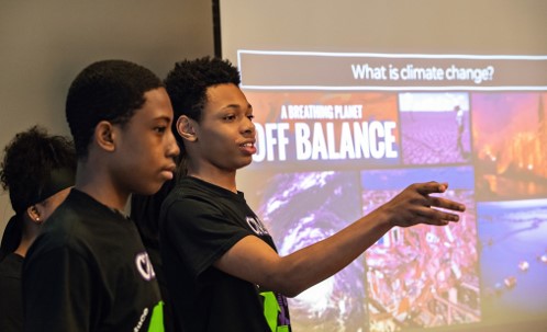 Three students in black shirts do a presentation on climate change they are standing in front of a large projector screen. One student is pointing into the crowd and smiling mid-sentance.