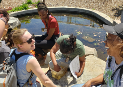 Two members are having a laugh while they practice water quality assessment at the DNR's Outdoor Adventure Center's pond areas along with Detroit River. Others are seated along the side of a pond in the sun.