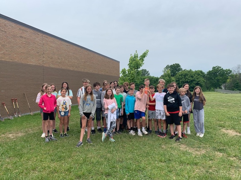 A Maltby 6th grade class of students stands in front of a newly planted tree in front of the school. Several students are holding tools and there are also some shovels lined up against the wall of the brick school.