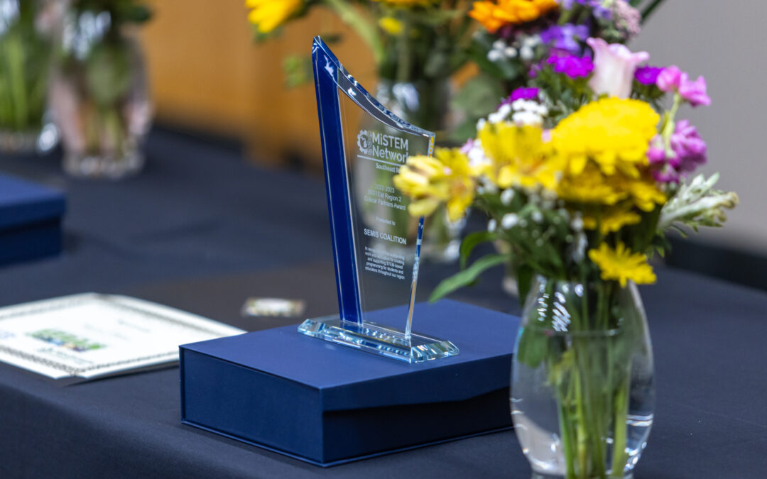 A photo of the MiSTEM Critical Partner award. There is a vase with yellow flowers not he right side of the image. The award is blue and clear. It sits on a blue box on a blue tablecloth. There are various colors of flowers in the background