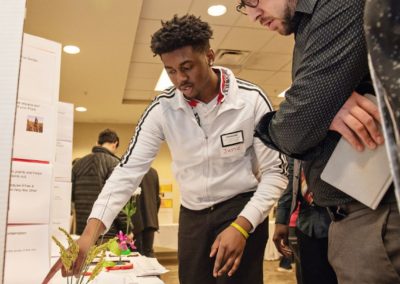 At the 2019 community forum a high school student wearing a white zip up points at a display board for the benefits of rain gardens while speaking to an older gentleman.