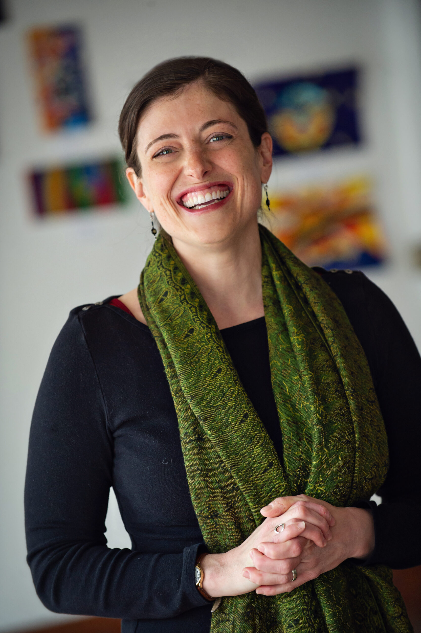 A woman smiles at the camera. She's wearing a black shirt with a green shirt and is clasping her hands in front of her. There is art on the walls in the background.