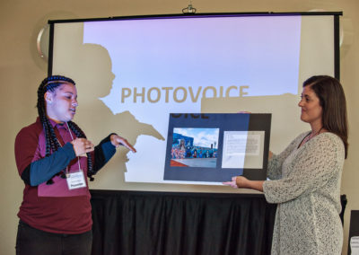 A student presents her photovoice project to the audience of the SEMIS Community Forum. She is wearing a dark red polo and looking to the right at a poster board that Lisa Voelker is holding to display for her.