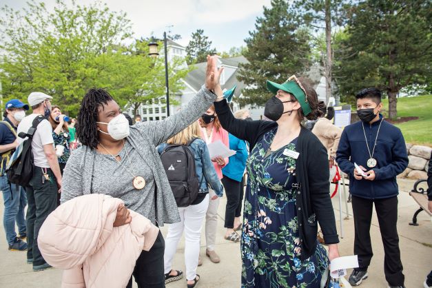 Lisa Voelker, SEMIS Assistant Director, wearing a flowery dress, a Black cardigan, and a green visor, is giving a high five to Latia Leonard, SEMIS community partner, wearing Black pants, a gray twin-set, and holding a rose jacket. They are outside near Eastern Michigan University Lake House during the May 19th Community Forum, with several people and trees behind them.