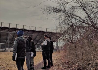 Community members are gathered for the YCHS Community walk in march 2022 looking up and left away from the camera. they are in cold weather wear. The sky is grey and there are leaves on the ground. in the background there is a stadium.