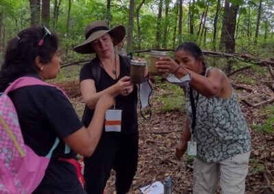 Three educators are holding up jars filled with water and soil in them. they are looking at soil samples in a forested space and discussing their findings amongst each other.