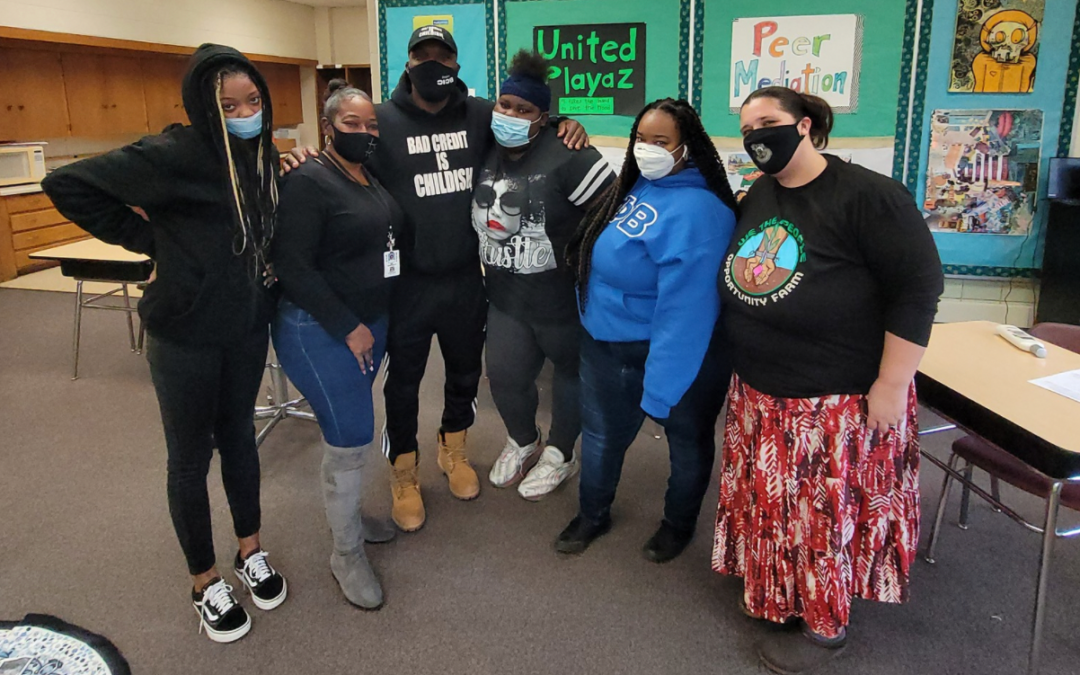 masked students and educators stand together and pose in front of a bulletin board located in a school classroom.