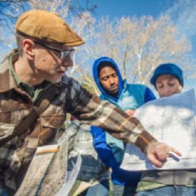 SEMIS member, historian and cultural landscapes expert Matt Siegfried wearing a brown flannel looking off to the right side at a map that is being held by two students. They are observing him point to an area of the map.