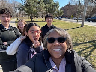 A selfie photo with six people in it. The person who took the photo Is wearing a black jacket with black sunglasses and is smiling. There Is grass and trees in the background.