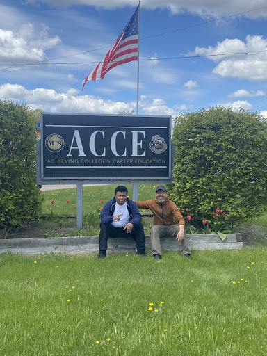 Mr. Pedro Betancourt (L) and Mr. David Vidal-Jones ( R) -  (ACCE Staff Members) . These two people are sitting in front of the Acheiving College and Career Education sign with a bush on either side of them, in a grassy green field with yellow dandelions.